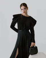 Woman posing with hand on hip holding a small bag, wearing a long dress and earrings.