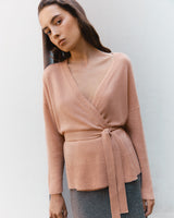 Woman standing in a V-neck sweater tied at the waist against a plain background.