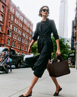 Person walking on city sidewalk carrying large bag, wearing sunglasses and jacket.