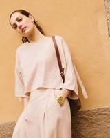 Woman in loose outfit leaning against a wall wearing a shoulder bag, large earrings and bracelet.