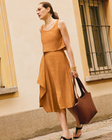 A person in a loose sleeveless top, and flowy skirt holds a large handbag walking by a building.