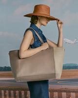 Person holding a large bag standing in front of a runway with a plane in the background, wearing a hat.
