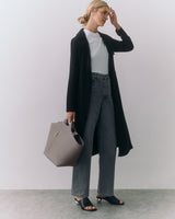 Person in long coat and jeans holding a large handbag standing indoors.