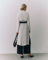 Woman in long coat and wide pants holds a handbag standing against plain background