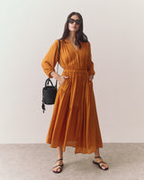 Woman in long dress with sunglasses holding a woven bag, standing indoors.