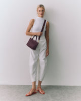 Person in sleeveless top and pants holding a handbag, standing against a plain wall.