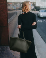Person standing on sidewalk holding a large bag with both arms crossed.