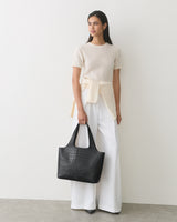 Model holding croc-embossed leather tote bag, wearing white pants and shirt.