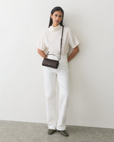 Person wearing a blouse and pants with a small shoulder bag, standing against a plain backdrop.