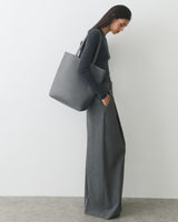 Person standing with large handbag and hands in pockets, against a plain background.