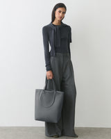 Person standing with a large tote bag, wearing a tied sweater and wide-leg pants.