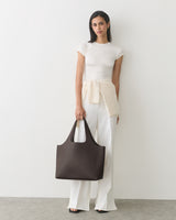 Person stands against a plain wall holding a large handbag and wearing a casual outfit.