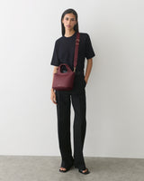 Person standing, holding a handbag with shoulder strap, wearing a plain outfit.
