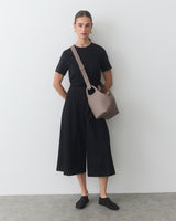 Person wearing a short-sleeve shirt, wide-leg pants, bag and slip-on shoes standing against wall