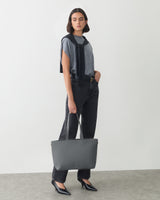 Person in casual outfit holding a large handbag, standing on a plain background.