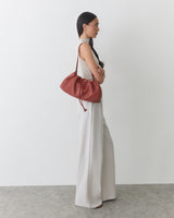 Person in sleeveless top and pants holding a shoulder bag standing sideways in a simple setting.