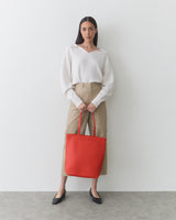 Person standing, holding a large tote bag with both hands, wearing long-sleeve top and pants.
