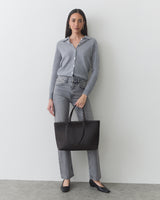 Person in casual outfit holding a tote bag standing against a plain backdrop.