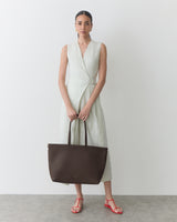 Person in sleeveless dress holding a large handbag, standing on a plain background.