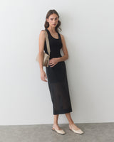 Woman in sleeveless dress standing with one hand on hip and holding a shoulder bag.