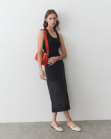 Woman in long dress with shoulder bag and slip-on shoes stands against a wall.