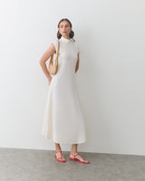Person wearing sleeveless dress with a shoulder bag and sandals in a minimal setting.