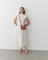 Woman in a dress with shoulder bag and sandals stands against a plain wall.
