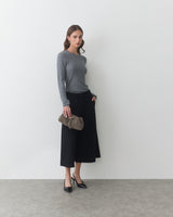 Woman standing in a gray top and black pants holding a clutch bag.