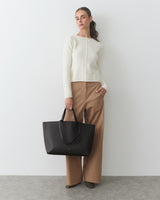 Person in sweater and wide pants holds large handbag, standing against plain background.