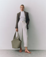Person in blazer and pants holding a bag, standing against a plain backdrop.