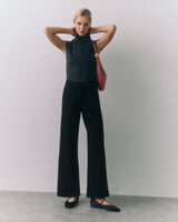 Person wearing sleeveless top, wide-leg pants, holding a bag, standing with arms raised.