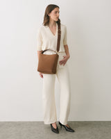 Person in casual outfit holding a shoulder bag wearing heels standing against a plain backdrop.
