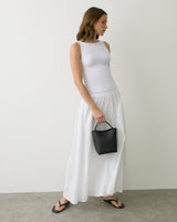 Person in long skirt and sleeveless top holds small handbag standing indoors.