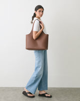 Person holding a tote bag, wearing jeans and sandals, standing against a plain background.