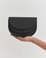 A hand holding a small, curved clutch bag against a plain background.