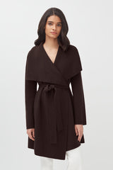 Woman in a belted coat standing against a plain background.
