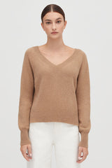 Woman in a V-neck sweater and white pants standing against a plain background.