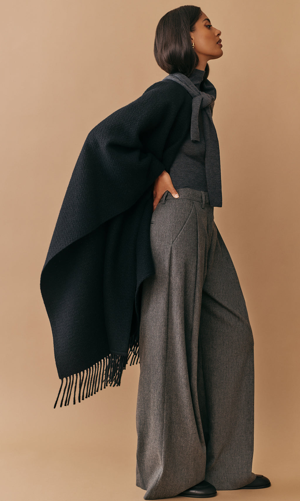 Person wearing a cape, scarf, and wide-leg pants standing sideways.