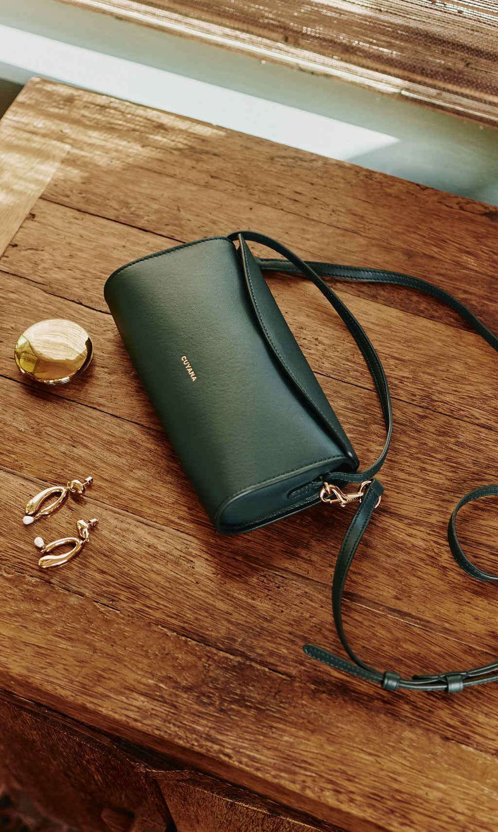 A handbag with a strap on a wooden surface next to earrings and a round compact.
