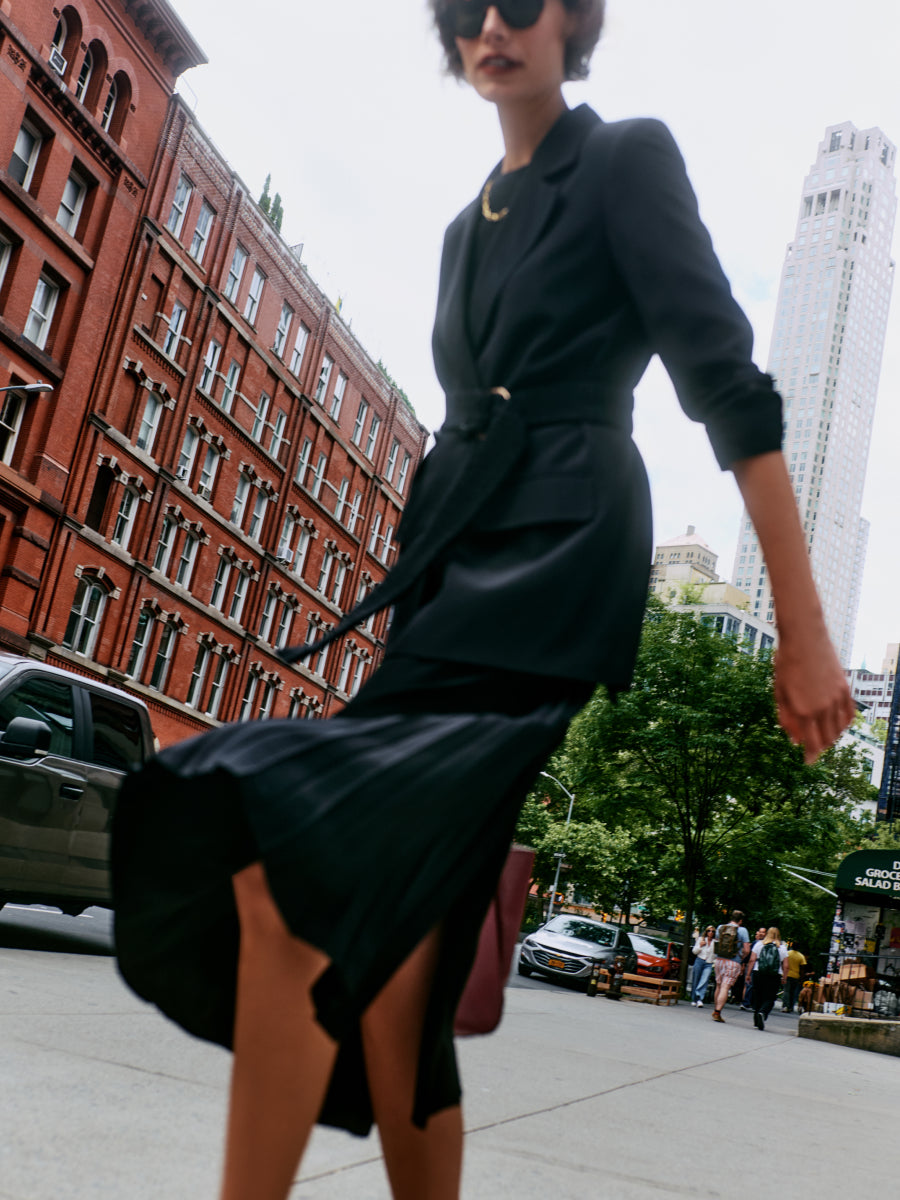 Person walking in city wearing belted blazer, skirt and sunglasses, carrying a bag