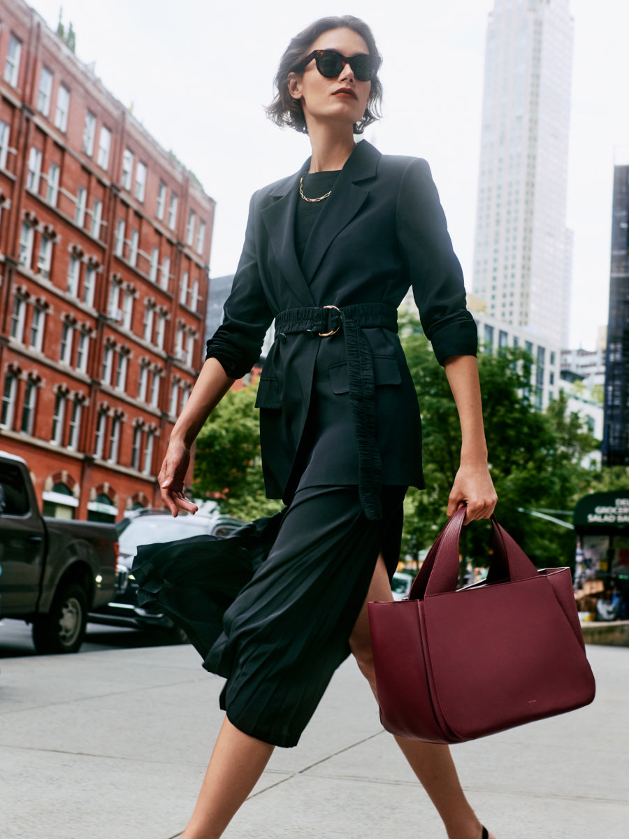 Person walking in city wearing belted blazer, skirt and sunglasses, carrying a large red bag