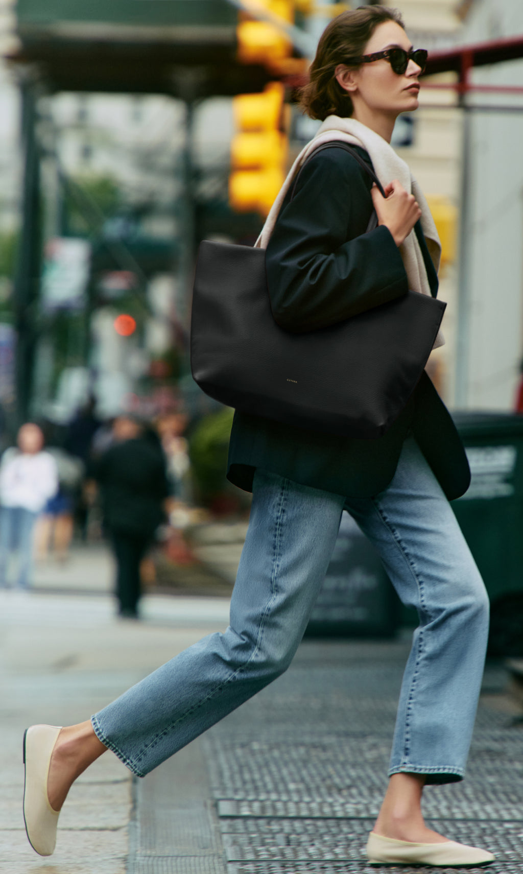 Person walking on sidewalk wearing sunglasses, carrying a large tote bag on shoulder.