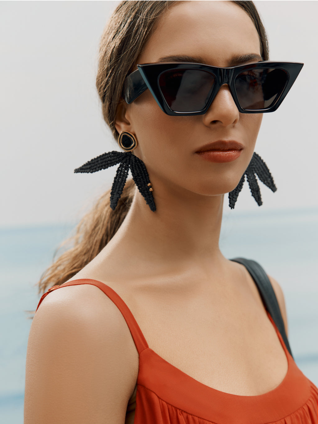 Woman wearing large sunglasses, leaf-shaped earrings, and a sleeveless top, standing outdoors