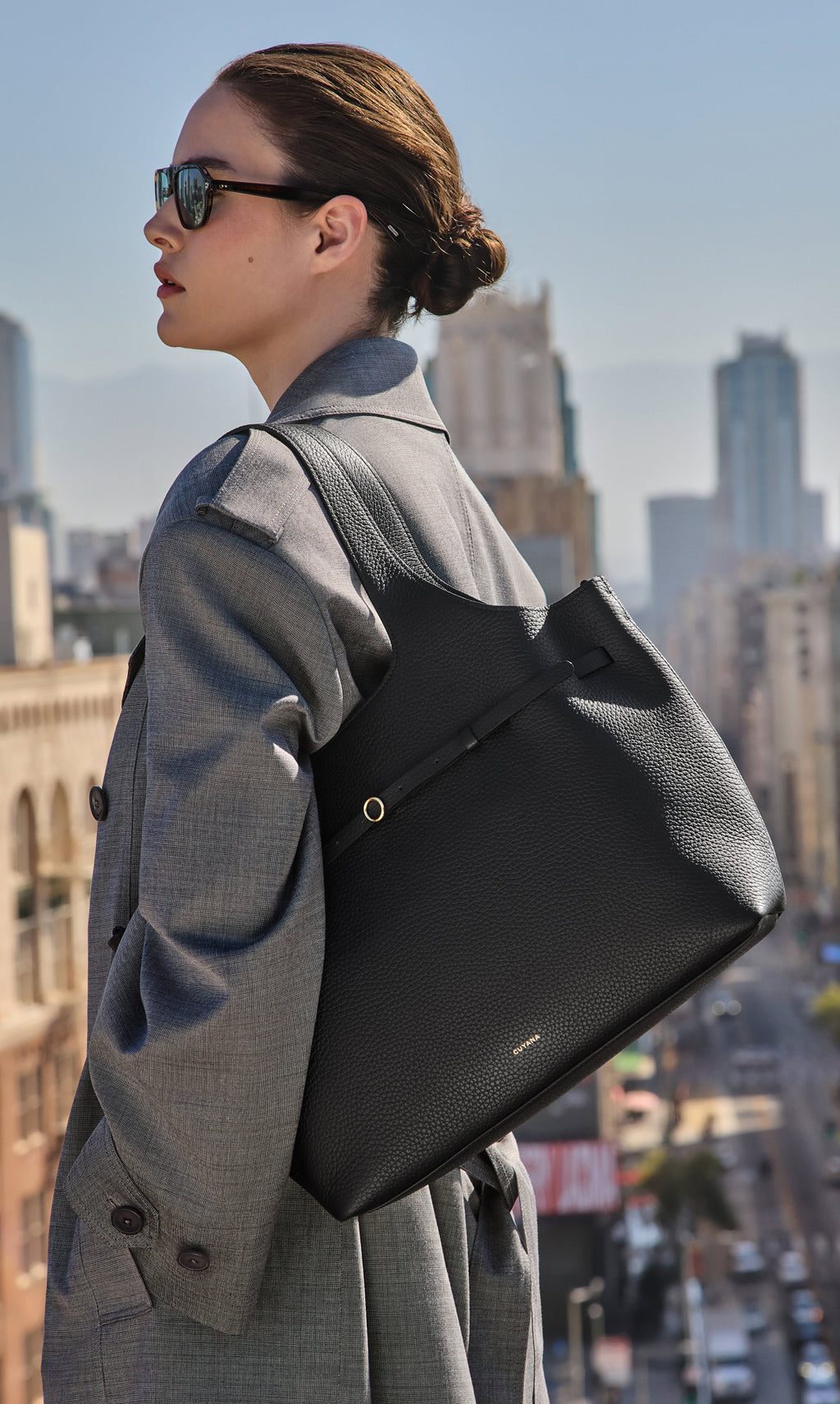 Person in a coat wearing sunglasses with a large handbag on a city rooftop.