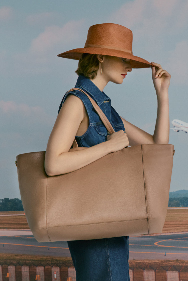 Woman holding her hat, carrying a large bag, near an airstrip.