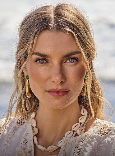 Person wearing shell necklace and earrings, standing outdoors with water in background.