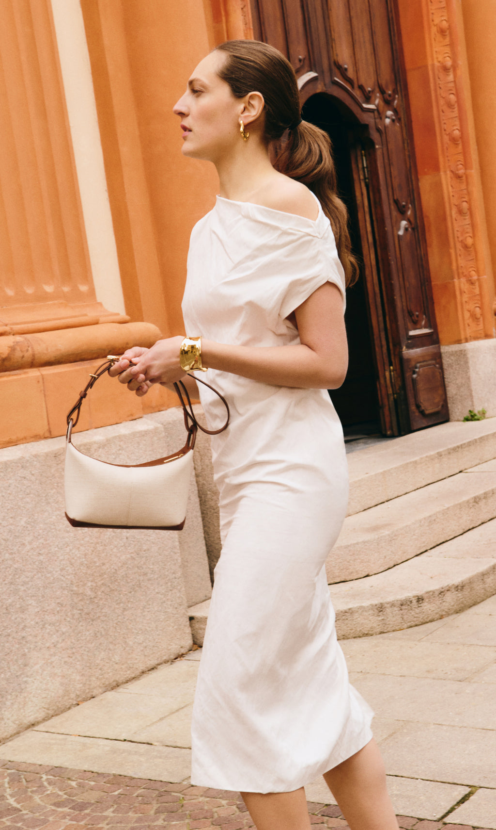 Woman in dress holding a handbag, walking near a building entrance.