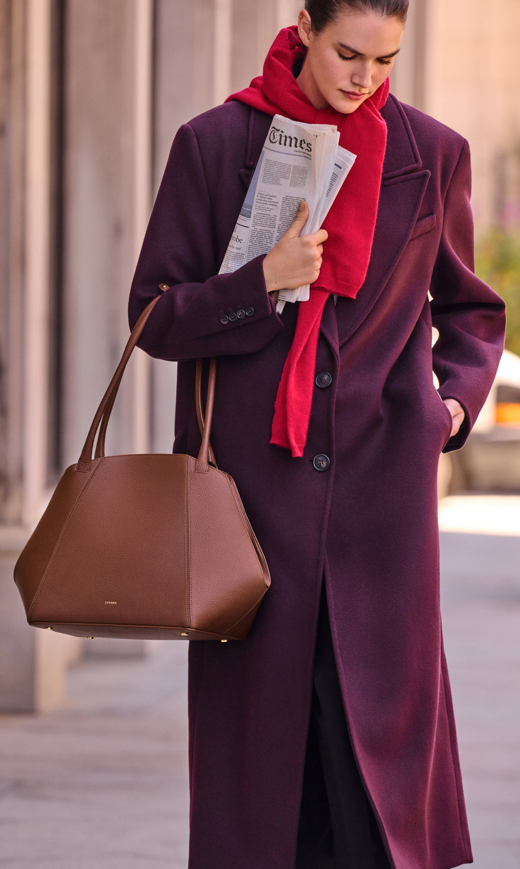 Person in coat with scarf holding newspaper and large handbag walking outside.
