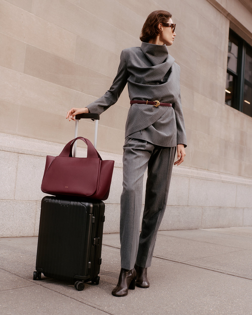Person standing with luggage and holding a large bag, wearing a belted suit and sunglasses