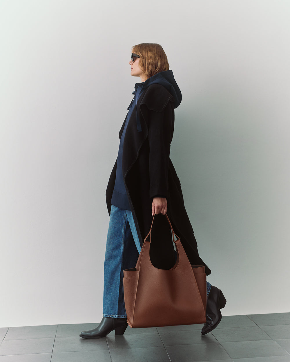 Person in a cape and jeans carrying a large bag, walking indoors.